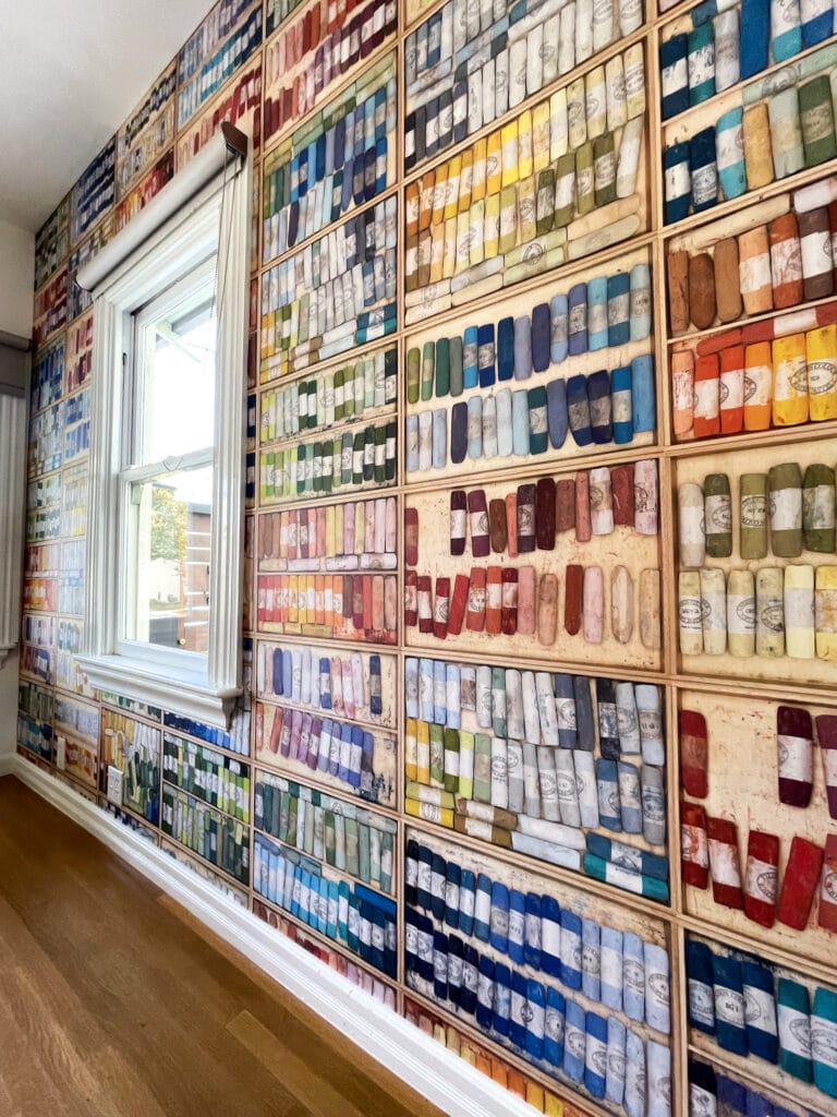 A wall covered in a wallpaper, expertly installed by a skilled wallpaper installer from the Bay Area, depicts numerous shelves filled with colorful tubes of paint. It’s positioned next to a white-framed window with blinds, and below, the light wooden floor is visible.