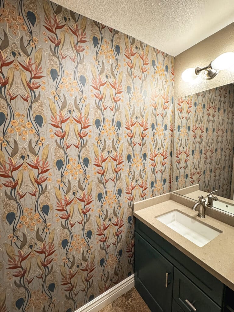 A bathroom with a beige countertop and dark green vanity. The walls, expertly adorned by a skilled wallpaper installation team from the Bay Area, feature a floral patterned wallpaper in shades of orange, blue, and gray. A two-light wall sconce is above the large mirror.