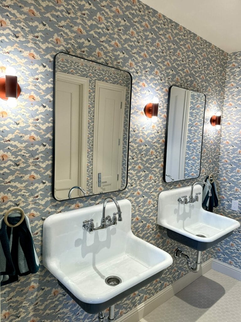 Two white sinks with mirrors on a patterned wallpapered wall, expertly accented by orange sconces. Dark hand towels hang beside each sink, creating a parfait of style and functionality.