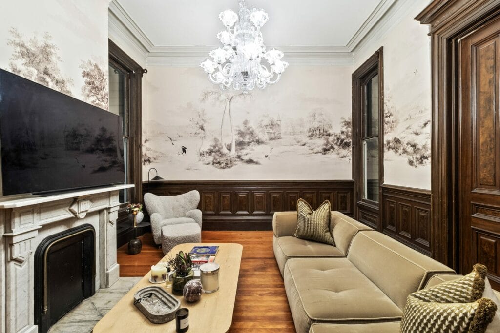 A living room with a beige sofa, armchair, and wooden coffee table sits under a luxurious chandelier. A wall-mounted TV complements the nature-themed wallpaper, expertly installed above dark wood panels, creating a parfait blend of elegance and tranquility.